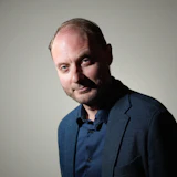 Professional headshot of a bearded man in blue blazer and navy shirt against neutral background