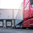 Red semi-truck backed up to warehouse loading dock with numbered bays during daytime