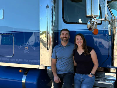 Two smiling people posing in front of a blue Tricycle Inc commercial truck on a sunny day