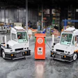 Two white industrial tug tractors flank a red electric charging station in a warehouse facility