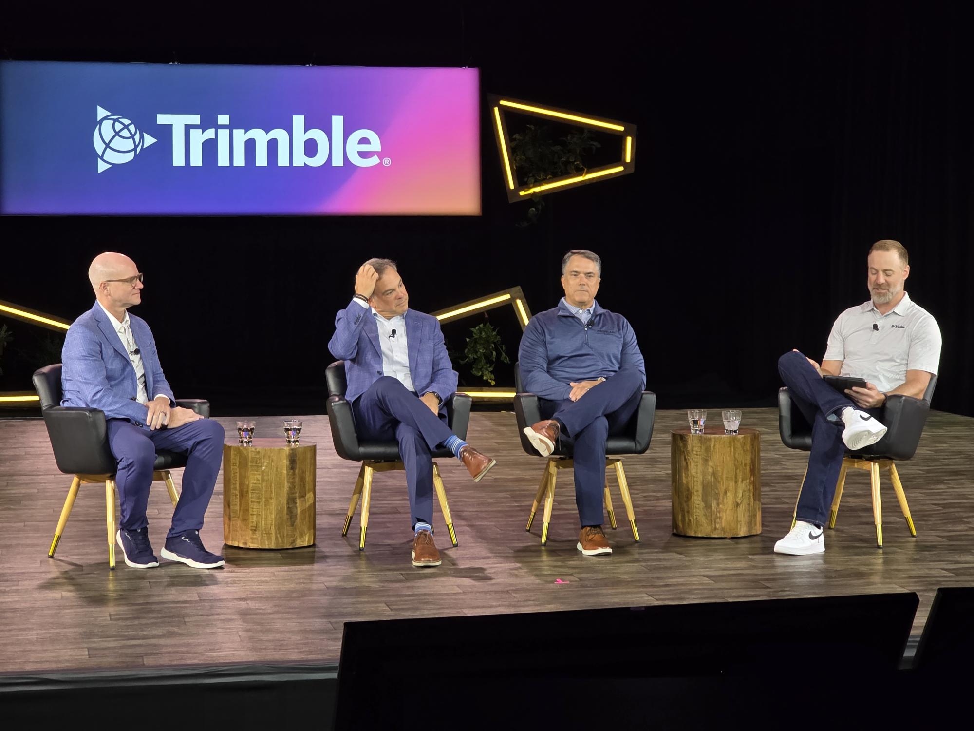Four business professionals participate in a panel discussion on stage beneath a Trimble company logo backdrop with yellow geometric design elements.