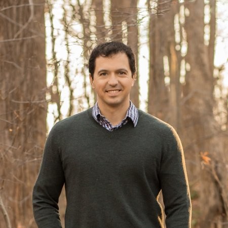 Smiling man in dark sweater and checkered shirt standing in a forest with bare trees Headshot