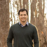 Smiling man in dark sweater and checkered shirt standing in a forest with bare trees