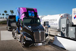 Black semi-truck with pink and blue custom sleeper cab parked among commercial vehicles and trailers in industrial lot