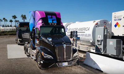 Black semi-truck with pink and blue custom sleeper cab parked among commercial vehicles and trailers in industrial lot