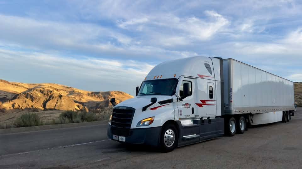 White and blue semi-truck with trailer parked on desert highway with rocky hills in background
