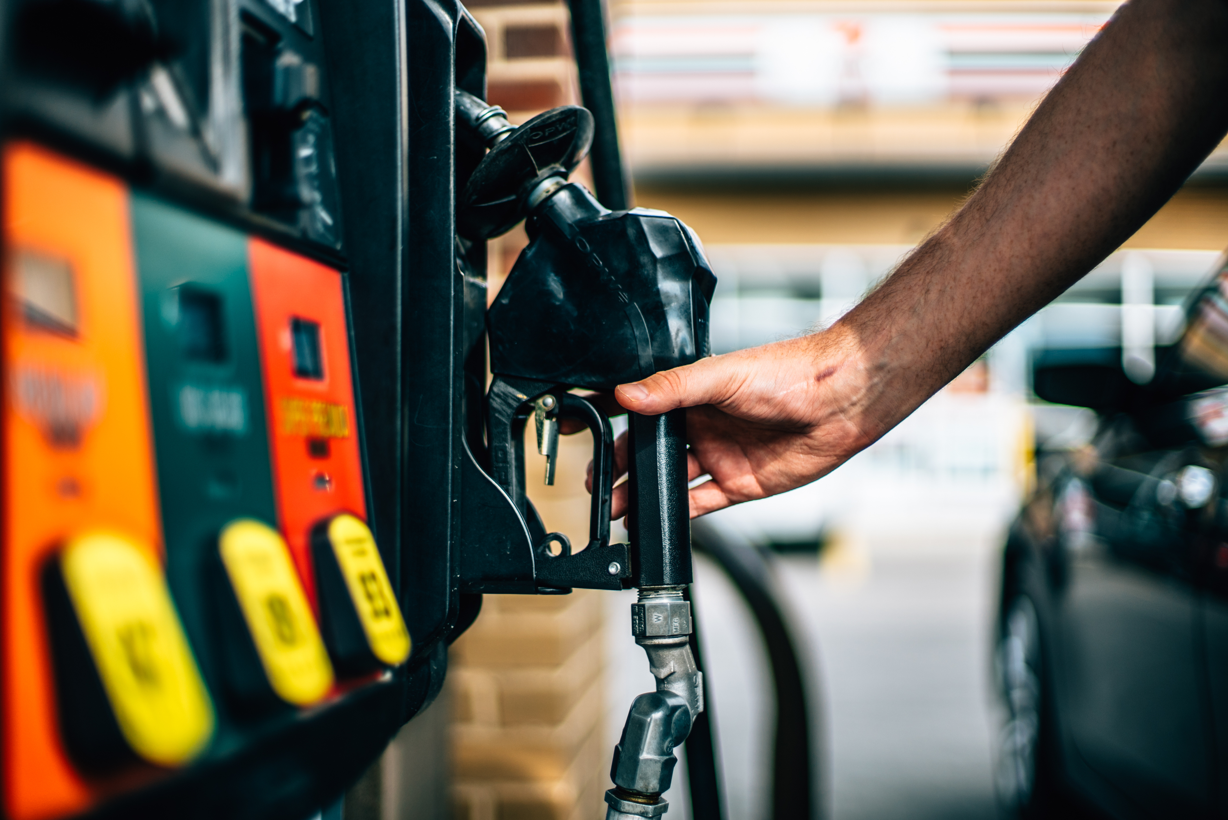 Person holding black fuel pump nozzle at gas station with orange and yellow pump interface visible