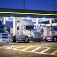 Fleet of commercial semi-trucks parked under illuminated canopy at truck stop during dusk