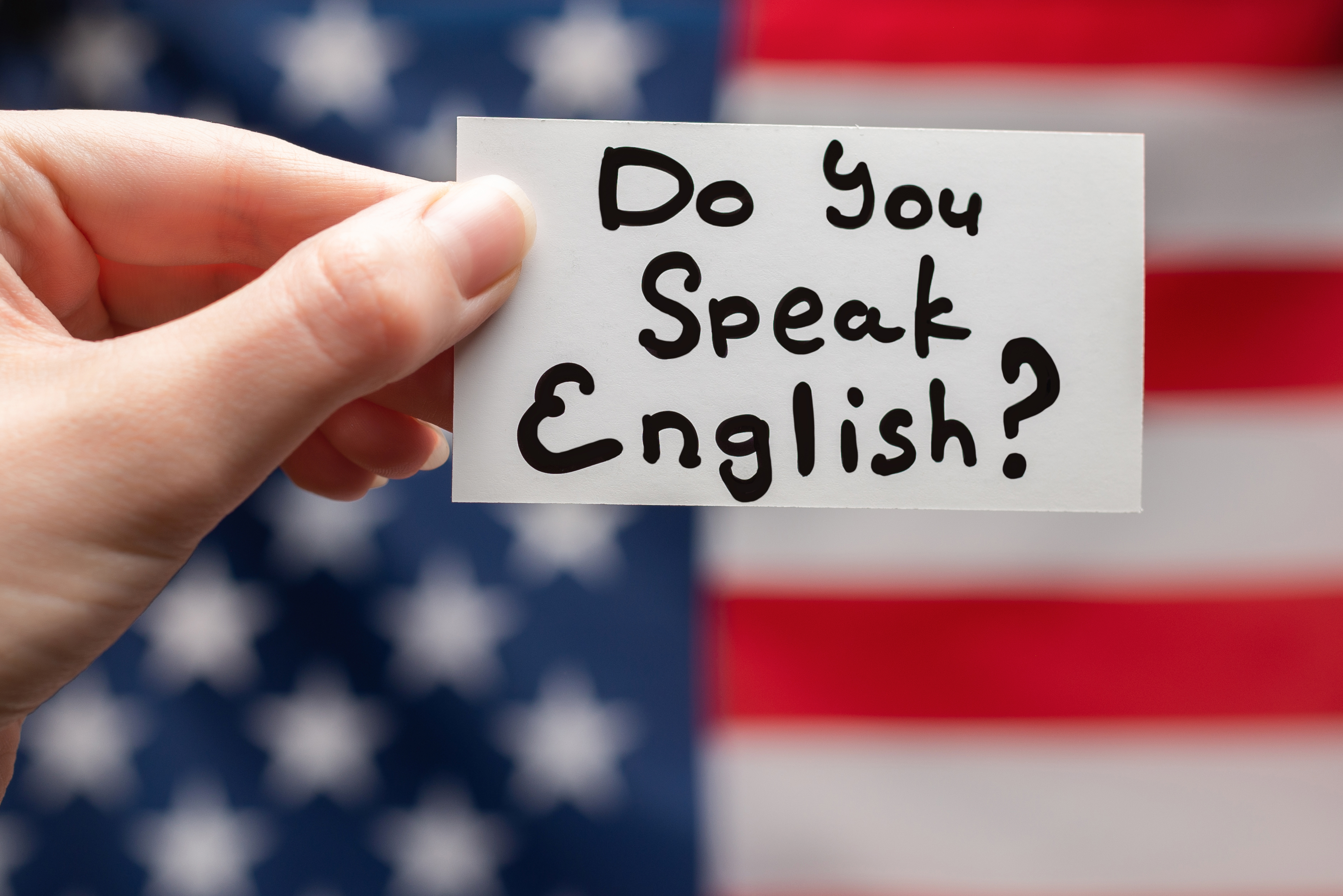 Hand holding white card with 'Do You Speak English?' written in black marker against blurred American flag background