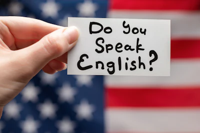 Hand holding white card with 'Do You Speak English?' written in black marker against blurred American flag background