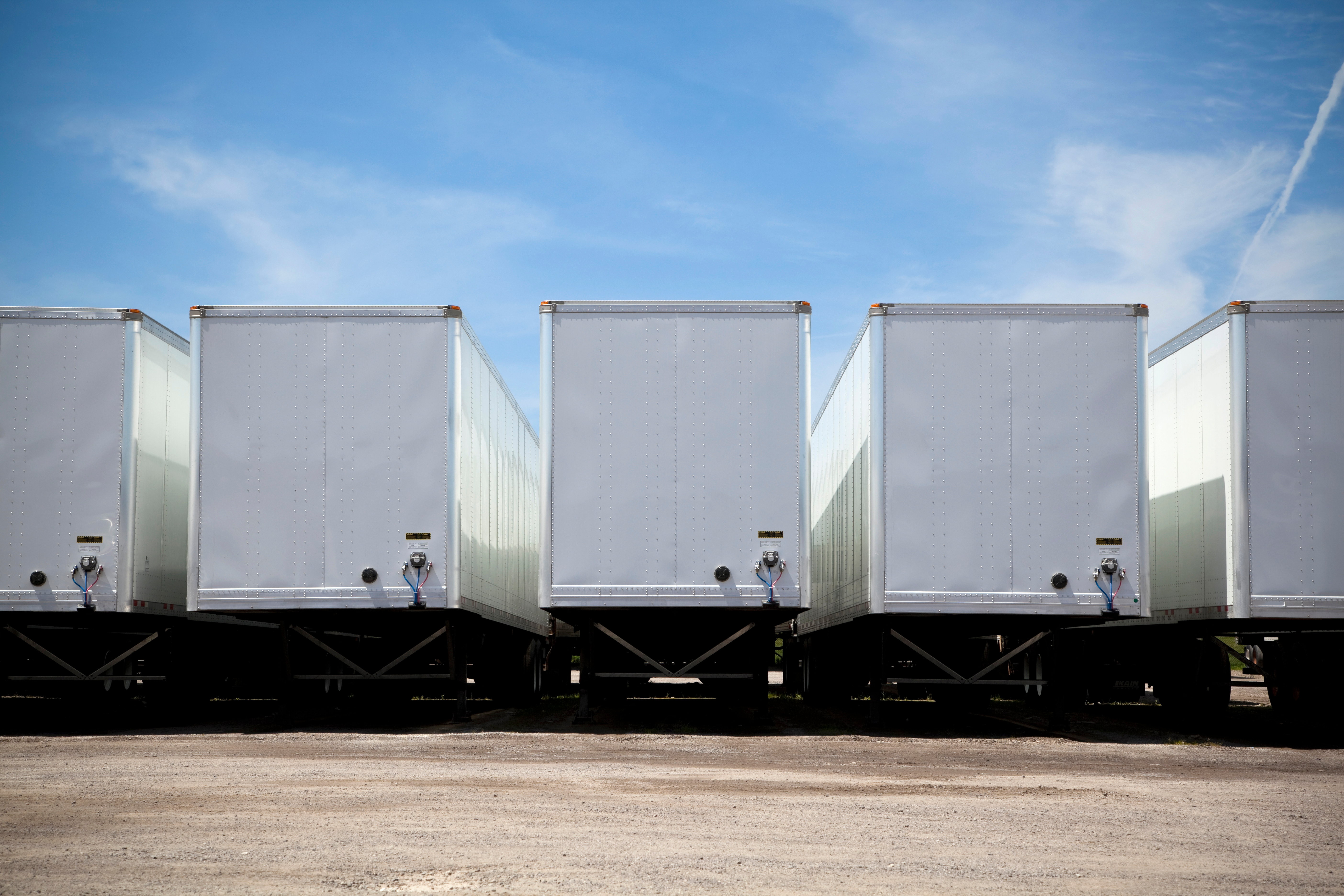 Row of white refrigerated trailers parked side by side on dirt ground under clear blue sky