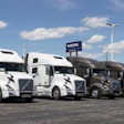 Row of white and brown Volvo semi-trucks parked at a dealership lot under blue sky