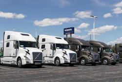 Row of white and brown Volvo semi-trucks parked at a dealership lot under blue sky