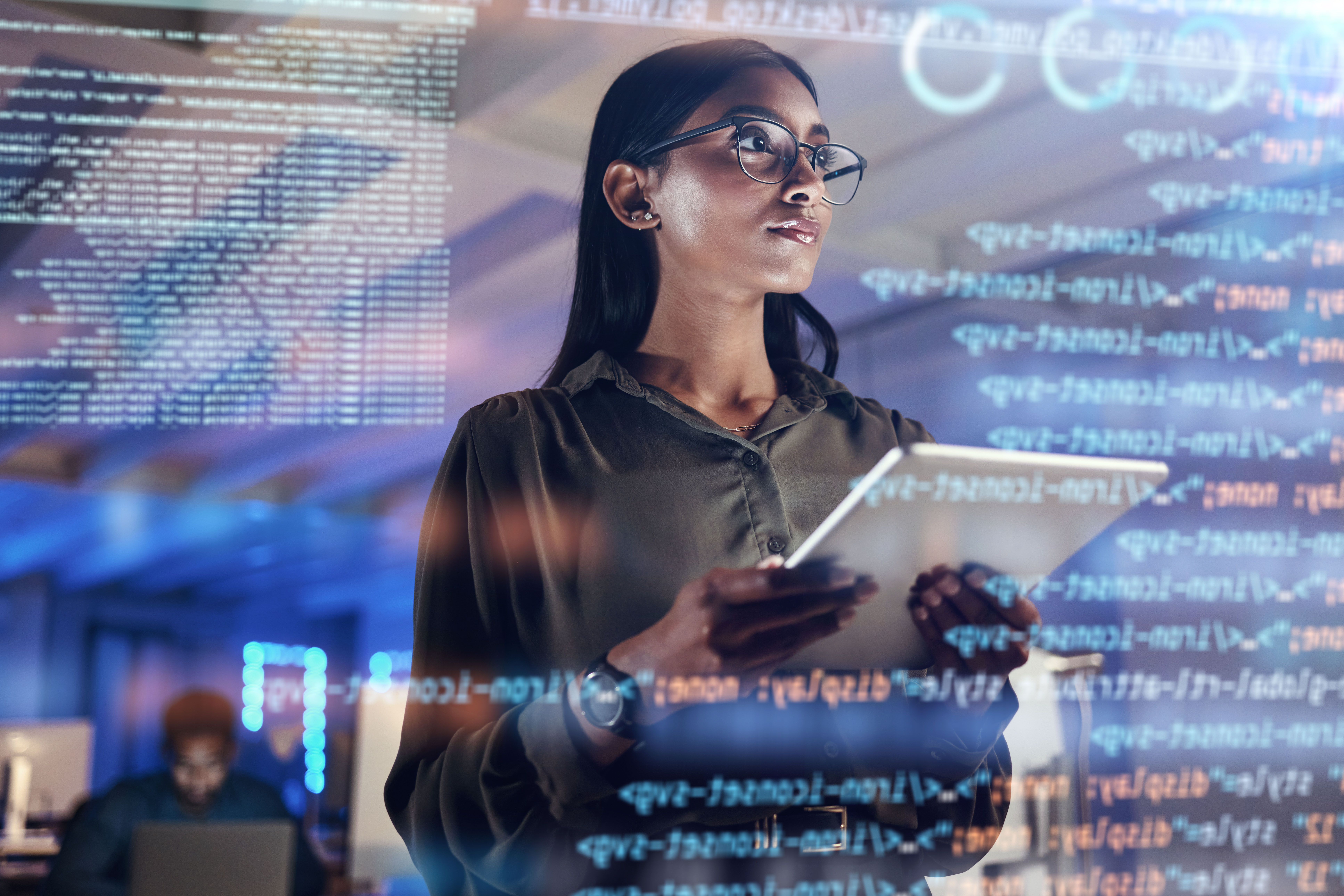 Female software developer wearing glasses holds tablet while examining code displayed on transparent screens in modern tech workspace