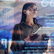 Female software developer wearing glasses holds tablet while examining code displayed on transparent screens in modern tech workspace