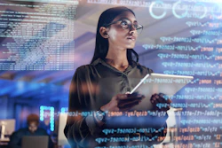 Female software developer wearing glasses holds tablet while examining code displayed on transparent screens in modern tech workspace