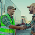 Two workers in safety gear shake hands in front of trucks at a logistics facility during sunset