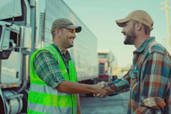 Two workers in safety gear shake hands in front of trucks at a logistics facility during sunset