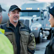 Three transportation workers smiling while discussing at a trucking facility with semi-trucks in the background