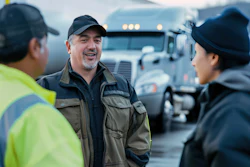 Three transportation workers smiling while discussing at a trucking facility with semi-trucks in the background
