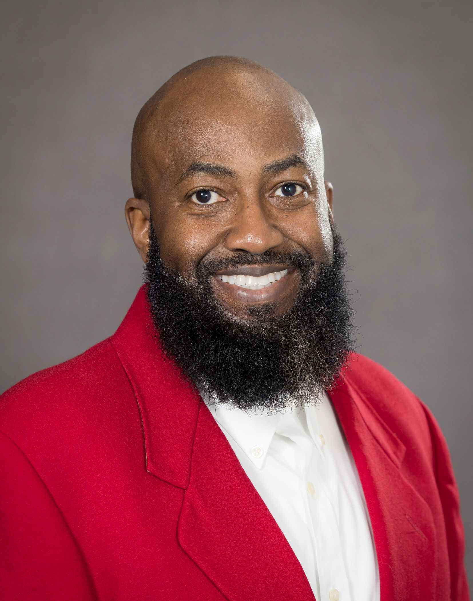 Professional headshot of a smiling man wearing a red blazer and white shirt against a gray background Headshot