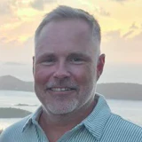 Professional headshot of a middle-aged man smiling at the camera with a coastal landscape background.