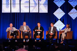 Five professionals participate in a panel discussion on stage with blue curtains and geometric background graphics