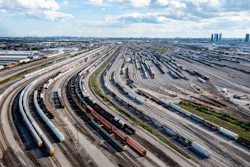 Aerial view of a sprawling railroad marshaling yard with multiple curved tracks and freight trains under a cloudy sky.