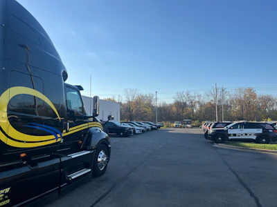 Black and yellow semi-truck parked in commercial lot with vehicles and bare trees under clear sky