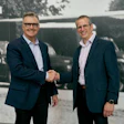 Two businessmen in navy blazers shake hands during a corporate meeting in front of a historical photograph backdrop