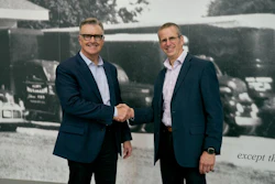 Two businessmen in navy blazers shake hands during a corporate meeting in front of a historical photograph backdrop