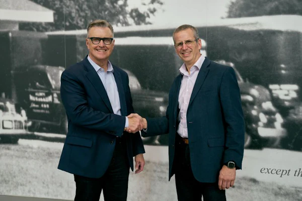 Two businessmen in navy blazers shake hands during a corporate meeting in front of a historical photograph backdrop