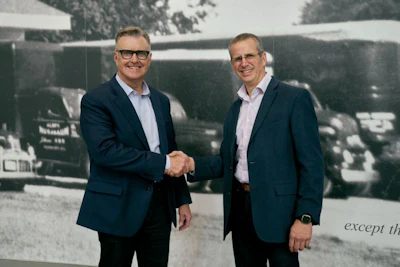 Two businessmen in navy blazers shake hands during a corporate meeting in front of a historical photograph backdrop