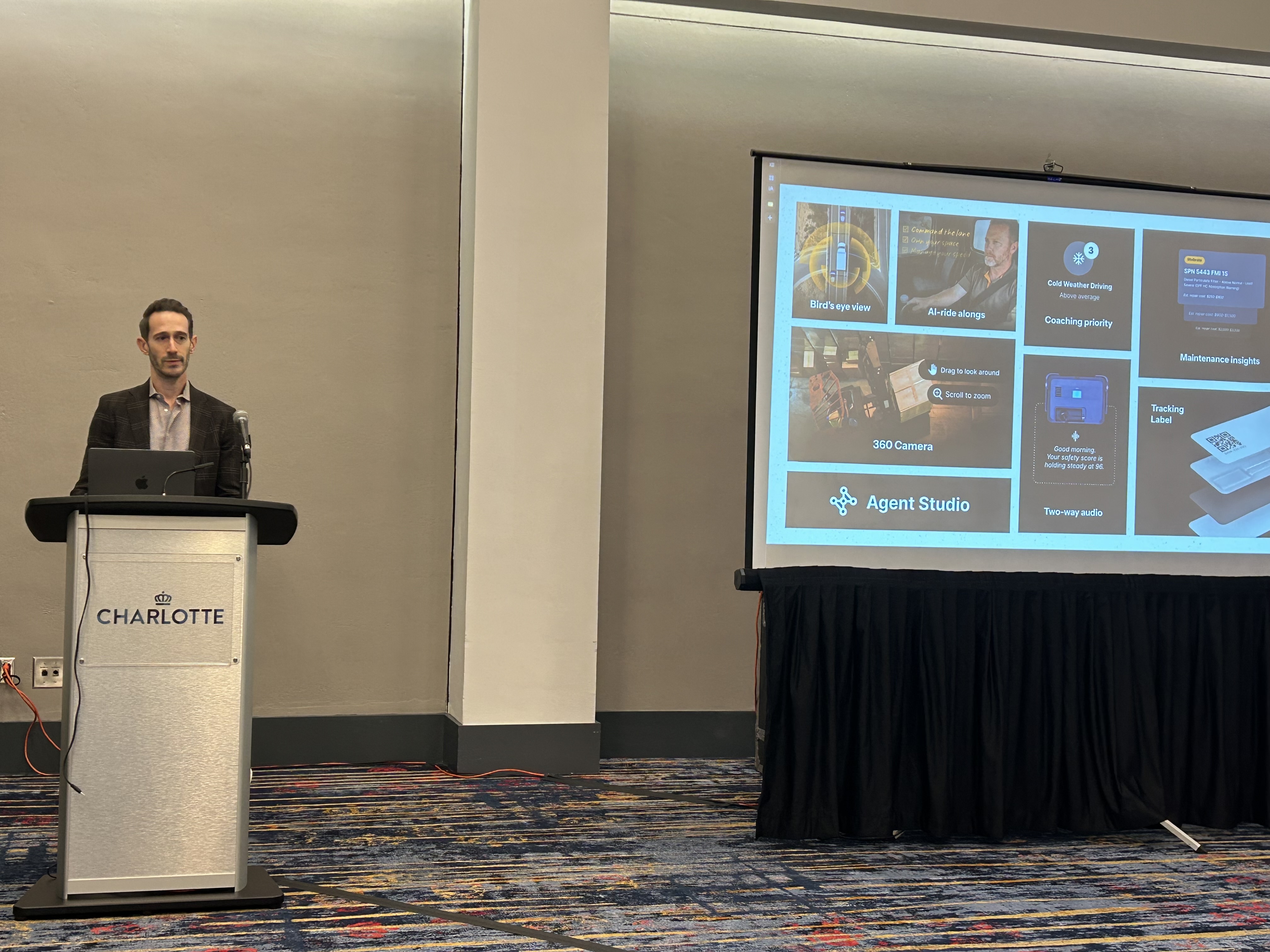 Man speaking at University of Charlotte branded podium during presentation with Agent Studio software interface displayed on screen