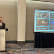 Man speaking at University of Charlotte branded podium during presentation with Agent Studio software interface displayed on screen