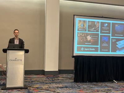 Man speaking at University of Charlotte branded podium during presentation with Agent Studio software interface displayed on screen
