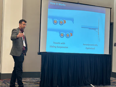Presenter in blazer speaking at conference with projection screen showing technical diagram of suspension system components