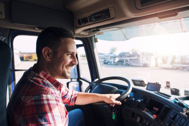 Smiling truck driver in red plaid shirt steering large commercial truck on highway