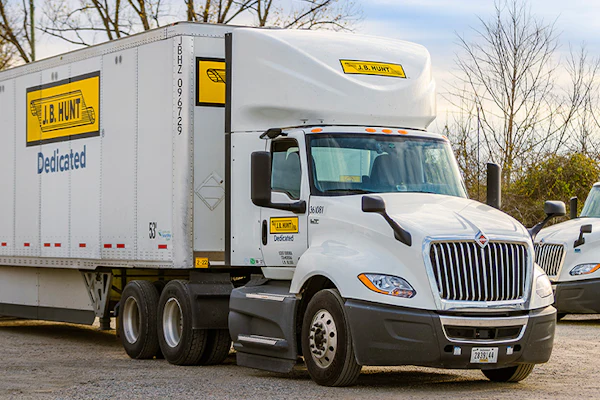 White J.B. Hunt semi-truck with yellow and black branding on the road with bare trees in background