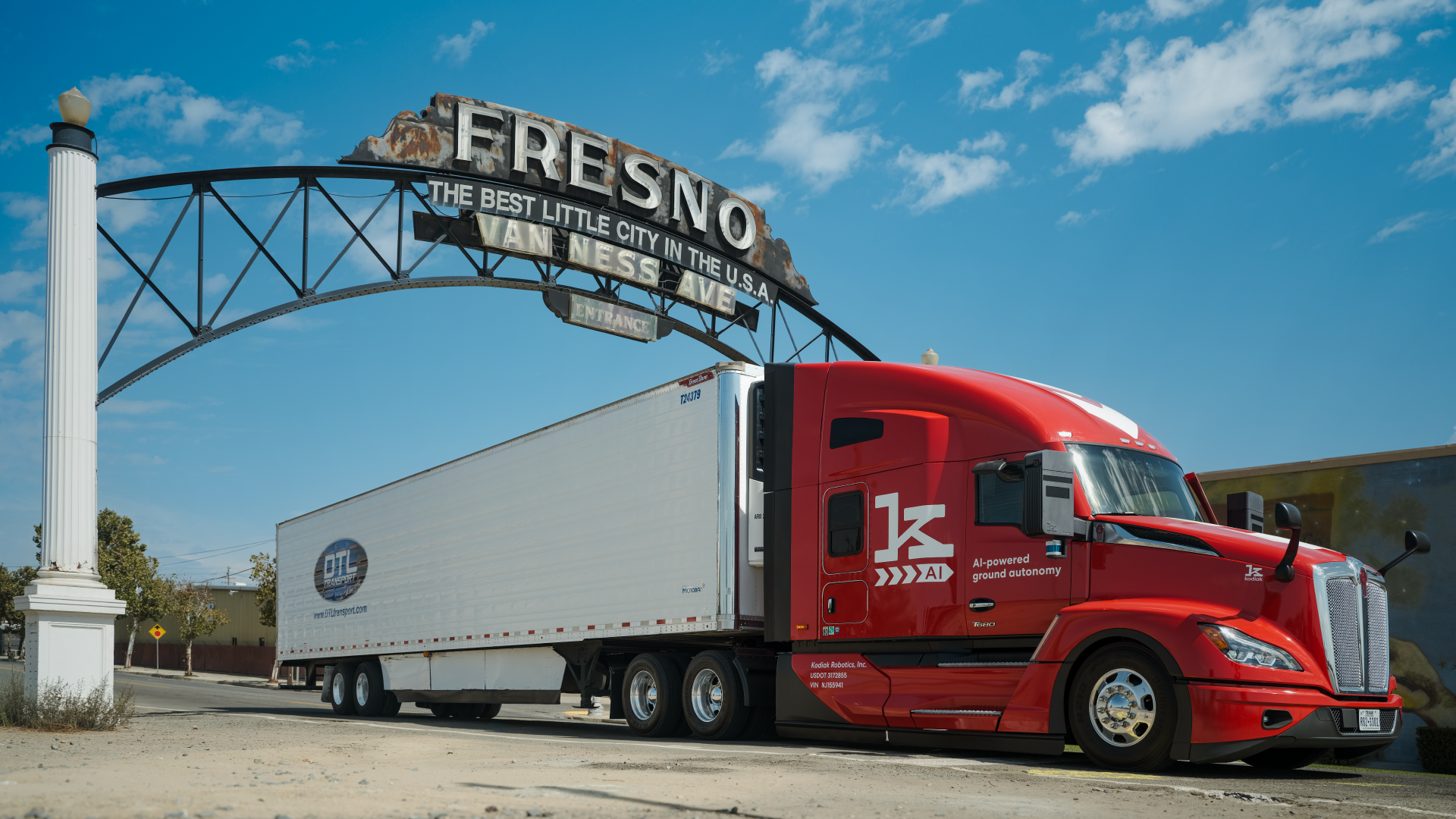 Red semi-truck with white trailer under Fresno's historic welcome sign against blue sky