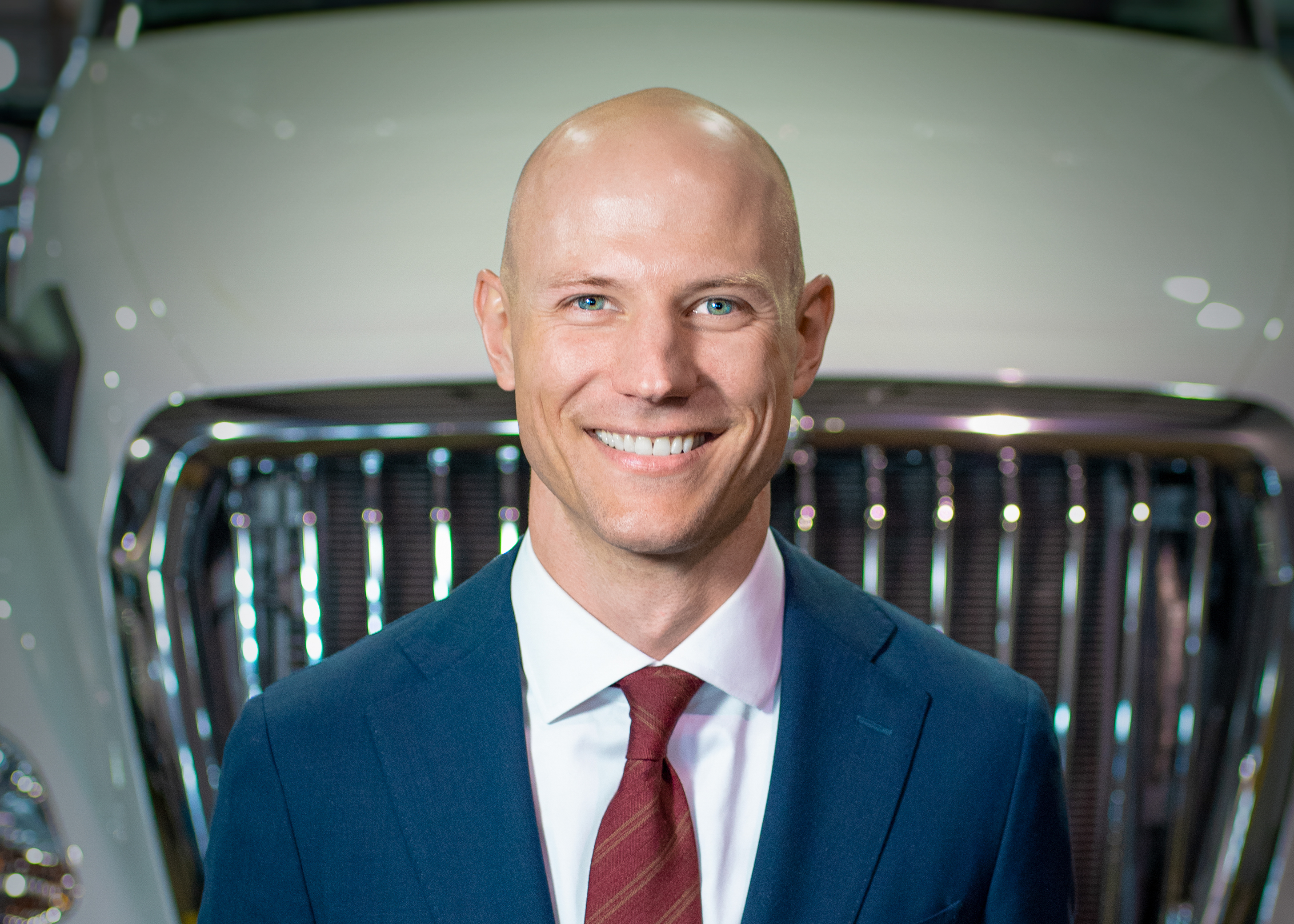 Professional headshot of smiling man in navy blazer and burgundy tie against vehicle background