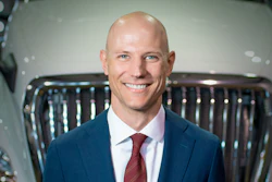 Professional headshot of smiling man in navy blazer and burgundy tie against vehicle background