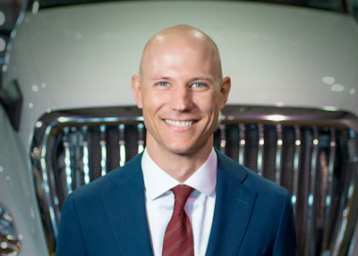 Professional headshot of smiling man in navy blazer and burgundy tie against vehicle background