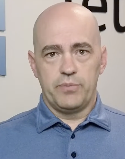 Bald man in blue button-up shirt against white background with partial text visible Headshot