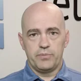 Bald man in blue button-up shirt against white background with partial text visible