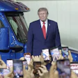 Man in navy suit standing beside blue truck at public event with crowd holding phones
