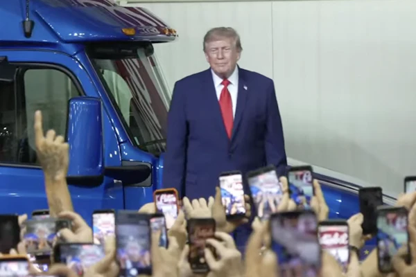 Man in navy suit standing beside blue truck at public event with crowd holding phones