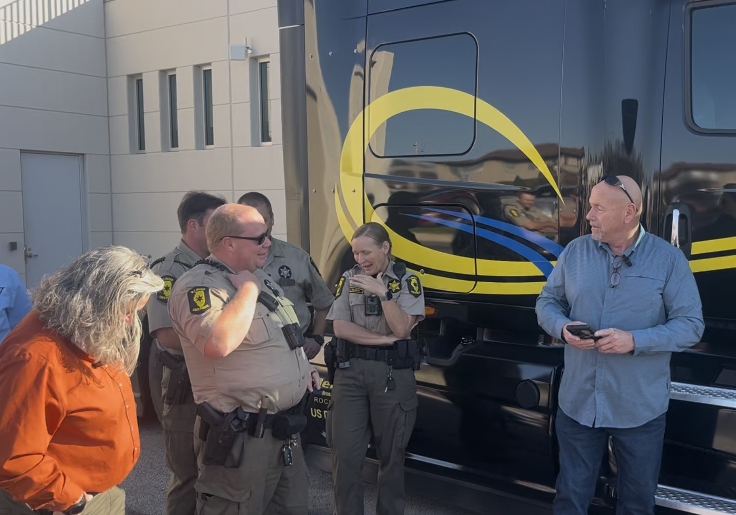 Law enforcement officers and civilians gathered near a yellow and blue striped bus during an outdoor community event.