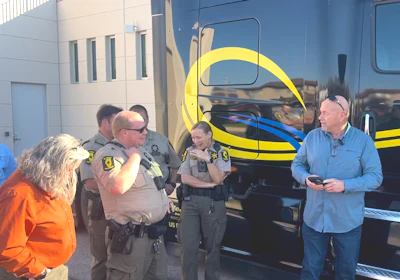 Law enforcement officers and civilians gathered near a yellow and blue striped bus during an outdoor community event.