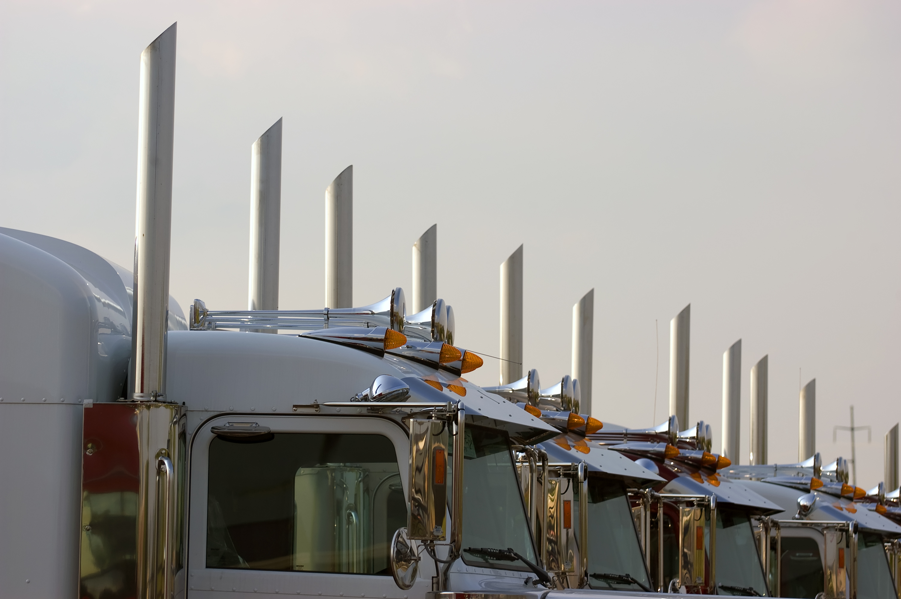 Fleet of semi-trucks lined up showing chrome exhaust stacks and cab designs against overcast sky